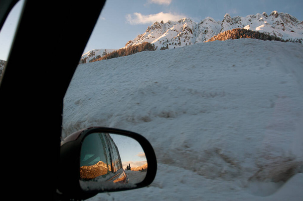 Vista nevada desde la ventana del coche. Puesta de sol en las montañas reflejo en el espejo retrovisor. Escape de invierno y concepto de viaje por carretera. Viajes locales. Italia, Dolomitas - Foto, imagen