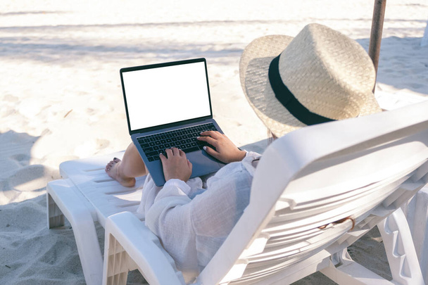 Mockup kép egy nőről, aki laptopot használ és gépel üres asztali képernyővel, miközben a strandszéken fekszik a strandon. - Fotó, kép