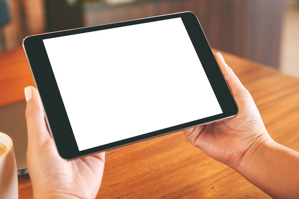 Imagen burlona de manos sosteniendo PC tableta negra con pantalla blanca en blanco con computadora portátil y taza de café en la mesa de madera - Foto, Imagen