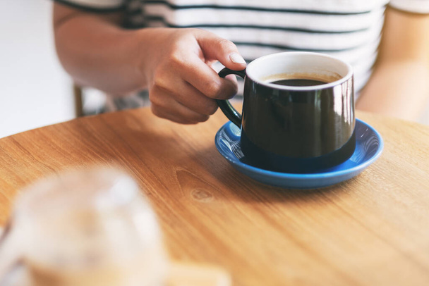 Imagen de cerca de la mano de una mujer sosteniendo una taza negra de café caliente sobre una mesa de madera - Foto, imagen