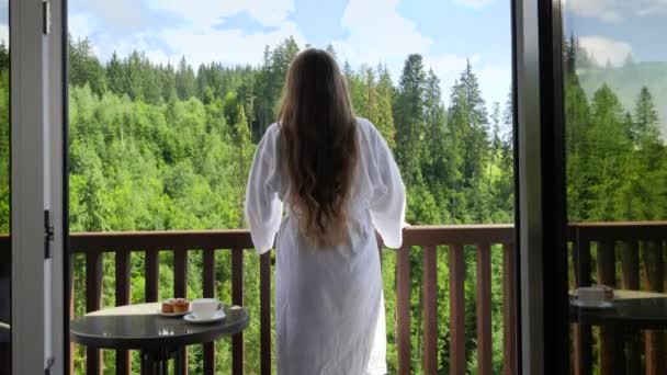 Vista posteriore di felice giovane donna in accappatoio allungando le mani dopo essersi svegliata al mattino in hotel di montagna. Concetto di turismo e vacanza in montagna in estate - Filmati, video