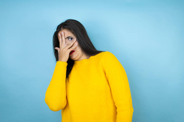 Młoda piękna kobieta ubrana w żółty sweter na odizolowanym niebieskim tle podglądająca w szoku twarz i oczy dłonią, patrząca przez palce ze wstydliwym wyrazem twarzy - Zdjęcie, obraz