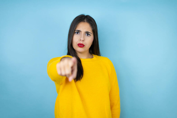 Young beautiful woman wearing yellow sweater over isolated blue background pointing with finger to the camera and to you, confident gesture looking serious - Photo, Image