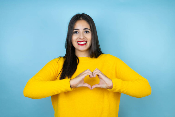 Молода красива жінка в жовтому светрі над ізольованим синім тлом посміхається в любові, показуючи символ серця і форму руками
 - Фото, зображення