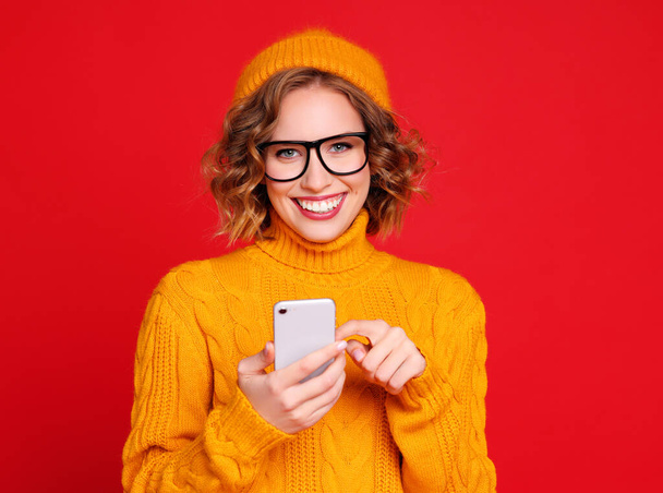 Aufgeregte trendige Frau lächelt und liest gute Nachrichten auf dem Smartphone und blickt vor rotem Hintergrund in die Kamera - Foto, Bild