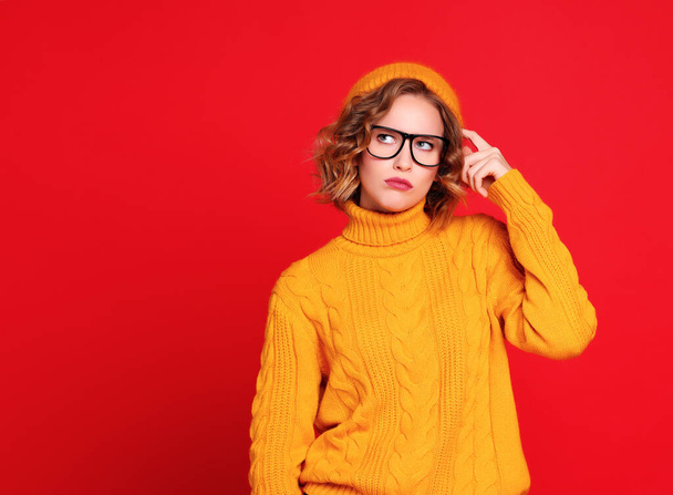 Pensive young woman in hat and glasses scratching head and looking up while thinking against red background - Photo, Image