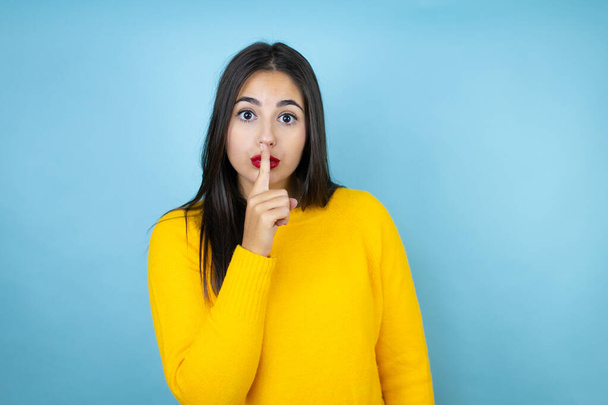 Giovane bella donna indossa maglione giallo su sfondo blu isolato chiedendo di essere tranquillo con il dito sulle labbra. Silenzio e concetto segreto. - Foto, immagini