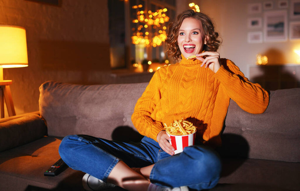 Mulher feliz em camisola de malha comendo batatas fritas e assistindo filme interessante enquanto ri e sentado de pernas cruzadas no sofá à noite em casa - Foto, Imagem
