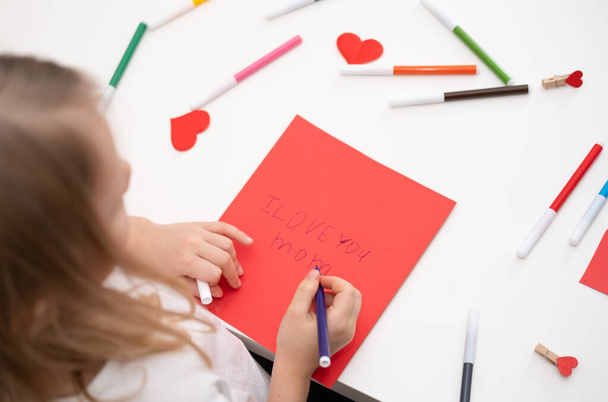 El niño dibuja una postal para la fiesta del Día de la Madre. Chica escribe en papel rojo Te amo mamá. - Foto, imagen