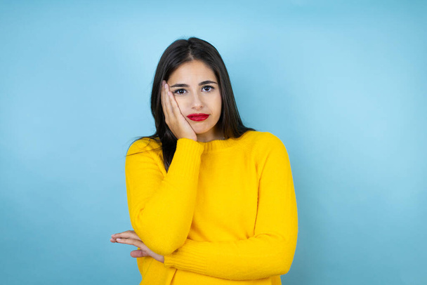 Młoda piękna kobieta ubrana w żółty sweter na odizolowanym niebieskim tle myśląc wyglądając na zmęczoną i znudzoną skrzyżowanymi ramionami - Zdjęcie, obraz
