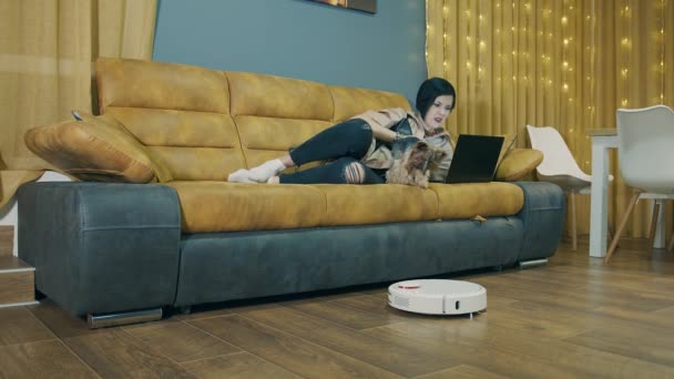 ソファに寝そべってノートパソコンで作業する女性と、ロボット掃除機が自動的に部屋を掃除します。自動ルームクリーニング。ペットと一緒に仕事をする自由時間. - 映像、動画