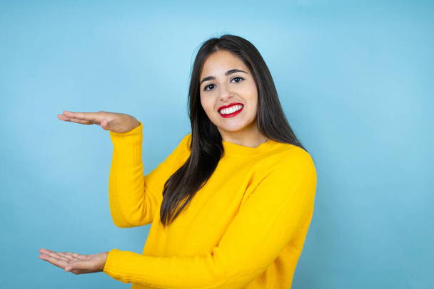Young beautiful woman wearing yellow sweater over isolated blue background gesturing with hands showing big and large size sign, measure symbol. Smiling . Measuring concept. - Photo, image