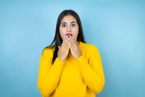 Young beautiful woman wearing yellow sweater over isolated blue background shocked covering mouth with hands for mistake. secret concept. - Photo, Image