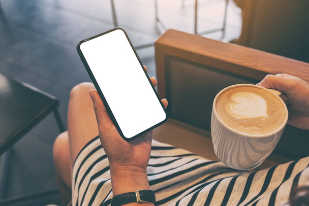 Imagen burlona de una mujer sosteniendo un teléfono móvil negro con pantalla en blanco mientras bebe café en la cafetería - Foto, Imagen