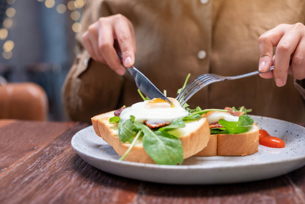 Uma mulher comendo sanduíche de café da manhã com ovos, bacon e creme de leite por faca e colher em um prato na mesa de madeira - Foto, Imagem