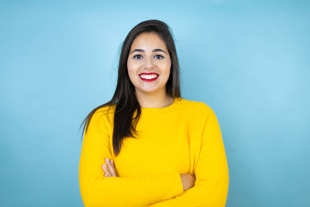 Junge schöne Frau in gelbem Pullover vor isoliertem blauen Hintergrund mit einem glücklichen Gesicht stehend und lächelnd mit einem selbstbewussten Lächeln, das Zähne mit verschränkten Armen zeigt - Foto, Bild