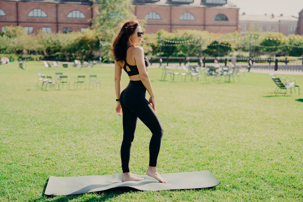 Spor kıyafetleri giymiş sağlıklı esmer kadın Karemat 'ın üzerinde çıplak ayakla duruyor yaz boyunca yeşil çimlerde incelik eğitimi alıyor güneş gözlüğü takıyor bir yerlerde fiziksel olarak iyi görünüyor. - Fotoğraf, Görsel