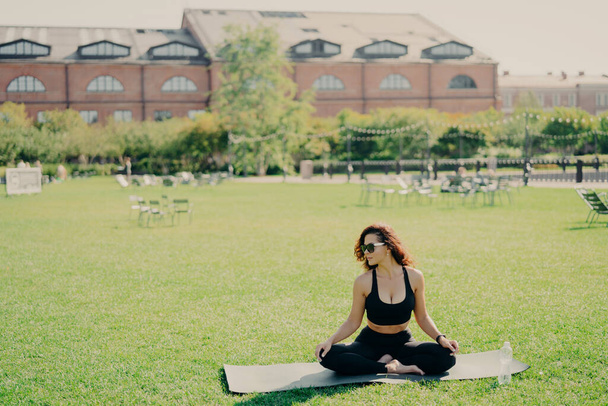Zrelaksowana szczupła brunetka Kaukaska kobieta siedzi w pozycji lotosu na macie fitness ma doskonały kształt ciała robi jogi i medytacji nosi ubrania sportowe stanowi na zewnątrz. Koncepcja kondycji zdrowotnej i relaksacji - Zdjęcie, obraz