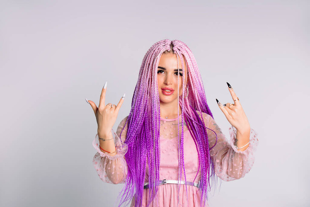 Young modern girl in a dress with long pink braids. Shows rock and roll gestures with both hands. White background. Side space. - Valokuva, kuva