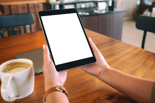 Mockup afbeelding van handen met zwarte tablet pc met blanco wit scherm met koffiekopje op houten tafel  - Foto, afbeelding