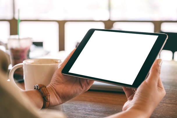 Obrázek rukou držících černý tablet s bílou obrazovkou vodorovně s šálkem kávy na dřevěném stole  - Fotografie, Obrázek