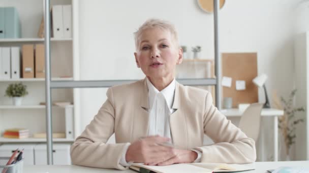 Rijpe blanke dame met kort grijs haar, in zakenpak en witte blouse aan het bureau met dagboek en pen in de hand en met iemand aan het praten - Video