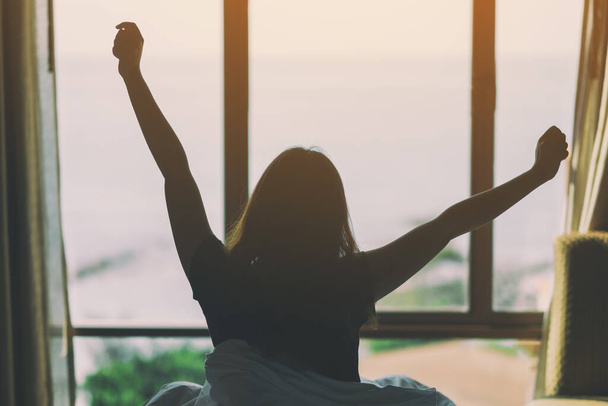 Женщина сидит и протягивает руки в кровати, глядя на красивый вид на море через окно после пробуждения утром - Фото, изображение