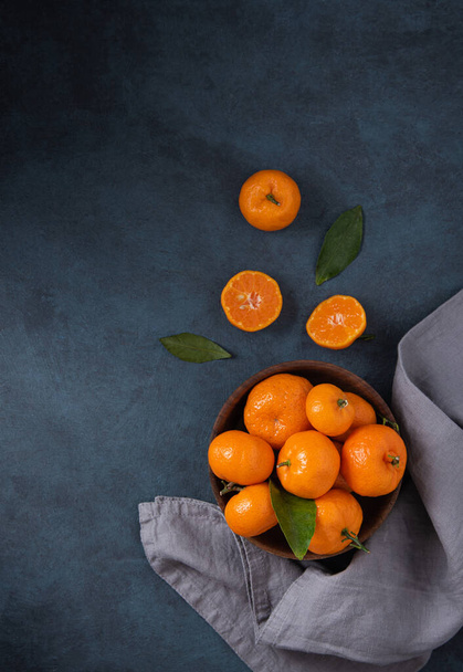 sweet tangerines with green leaves in wooden bowl  on a dark blue background with gray napkin. Dark photo. Top view and copy space - Φωτογραφία, εικόνα