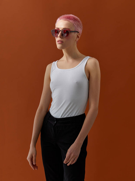 Chica Hipster con pelos cortos de color rosa, con camiseta blanca y jeans negros y gafas de sol con borde de madera y lentes rojas, sobre fondo marrón. - Foto, Imagen