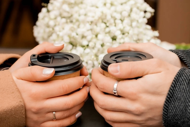 miesten ja naisten käsissä on kaksi ruskeaa lasillista kahvia, mustalaisen kukkakimpun vieressä - Valokuva, kuva