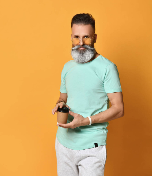 Moderní hipster muž drží jednorázové papírový kelímek, zatímco stojí na jasně oranžovém pozadí. - Fotografie, Obrázek