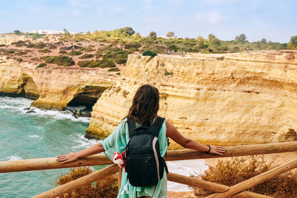 одна красивая сексуальная девушка стоит с большим рюкзаком у забора на скале в Португалии и смотрит вдаль, вид со спины на океан - Фото, изображение