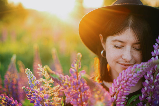 Portrait of beautiful woman holding lupine bouquet in sunset light in countryside field. Tranquil atmospheric moment. Young female with lupine flowers in warm sunshine in summer meadow - Фото, зображення