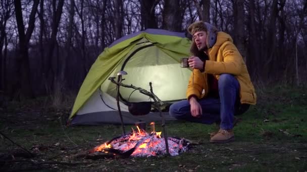 Un uomo beve tè vicino a un falò e una tenda nella foresta - Filmati, video