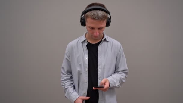 Ένας νεαρός άνδρας σε γκρι φόντο, με ακουστικά, πληκτρολογεί κείμενο σε ένα smartphone. Κοιτάζει προσεκτικά. - Πλάνα, βίντεο