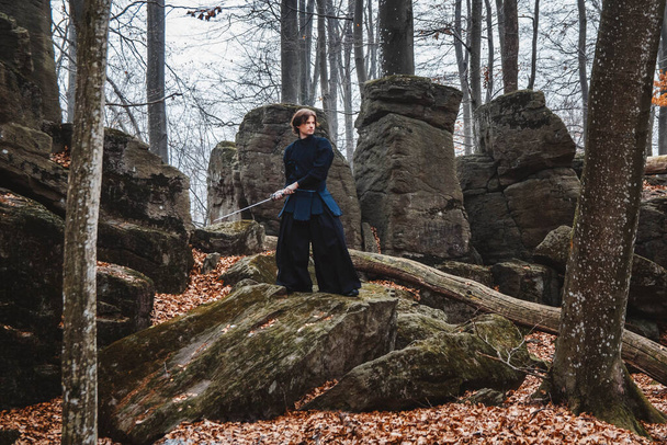 Joven en kimono negro practicando artes marciales con una espada sobre rocas y fondo del bosque. Lugar para texto o publicidad. - Foto, imagen