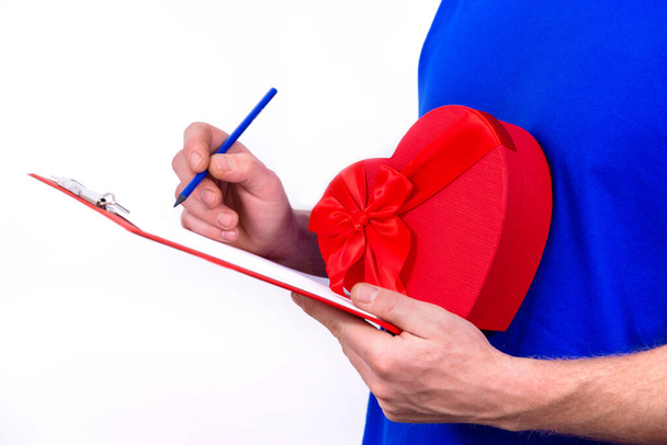 Courier in blue uniform holds red heart-shaped gift box for Valentines Day and checks order on clipboard. Home delivery gifts to your favorite women on Mothers and International Womens Day. - Foto, afbeelding