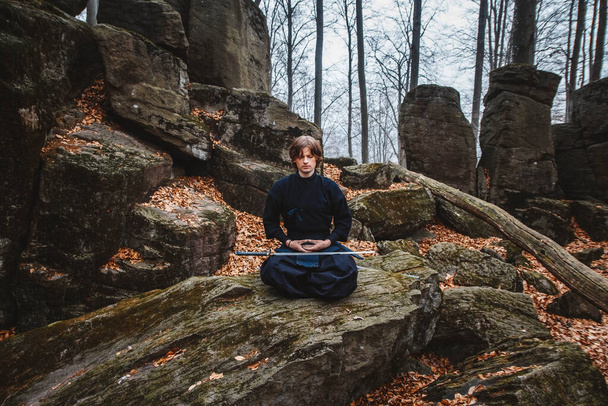 Młody człowiek w czarnym kimono z mieczem medytuje i koncentruje się na skałach i tle lasu. Miejsce na tekst lub reklamę. - Zdjęcie, obraz