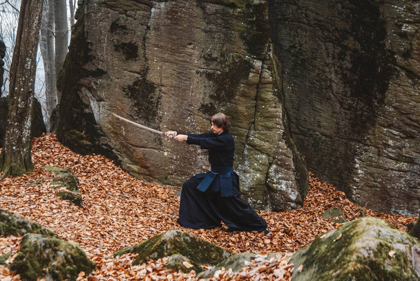 Jovem de kimono preto praticando artes marciais com uma espada sobre rochas e fundo da floresta. Lugar para texto ou publicidade. - Foto, Imagem