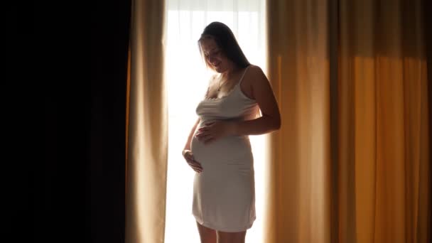 Silhouet van een mooie glimlachende zwangere vrouw die voor een groot raam in de slaapkamer staat. Concept van gelukkige zwangerschap en het verwachten van een baby - Video