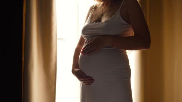 Close-up de bela mulher grávida em vestido de noite em pé contra o sol brilhante brilhando através de grande janela e acariciando sua barriga grande. Conceito de gravidez feliz e esperando um bebê - Filmagem, Vídeo