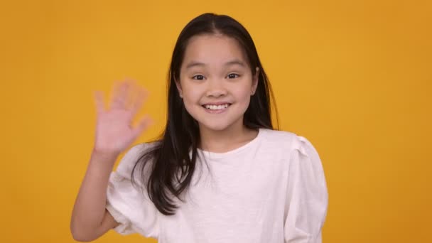 Cute friendly asian girl waving hand hi and smiling to camera, orange studio background - Footage, Video