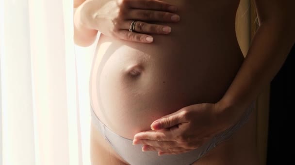 Gros plan de la femme enceinte touchant doucement et caressant son ventre en croissance à côté d'une grande fenêtre dans la chambre à coucher à la maison. Concept de grossesse heureuse et anticipation du bébé - Séquence, vidéo