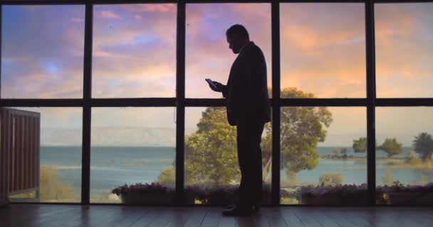 Geschäftsmann im Anzug checkt sein Telefon, während er bei Sonnenuntergang vor einem großen Fenster steht. Zeitlupe, Weitschuss. - Filmmaterial, Video