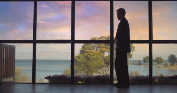 Liikemies kävelee lähellä suurta ikkunaa auringonlaskun aikaan, odottaen jotakuta, katsellen järvelle. Hidas liike, kaukaa haettu. - Materiaali, video