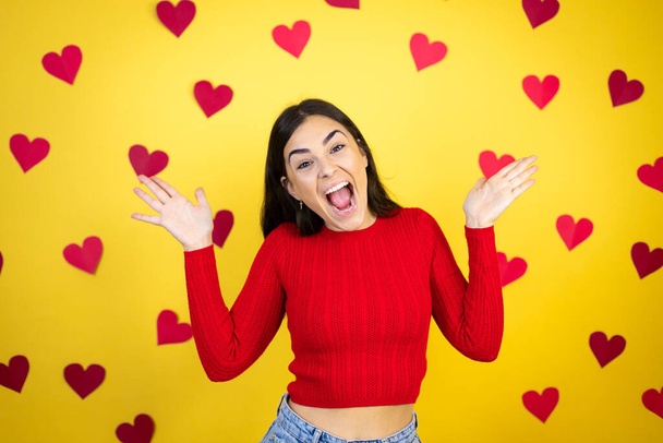 Young caucasian woman over yellow background with red hearts celebrating crazy and amazed for success with arms raised and open eyes screaming excited.winner concept - Foto, immagini