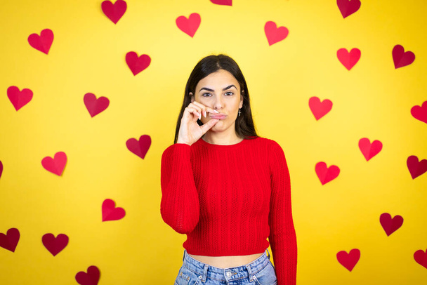 Young caucasian woman over yellow background with red hearts mouth and lips shut as zip with fingers. Secret and silent, taboo talking - Photo, Image