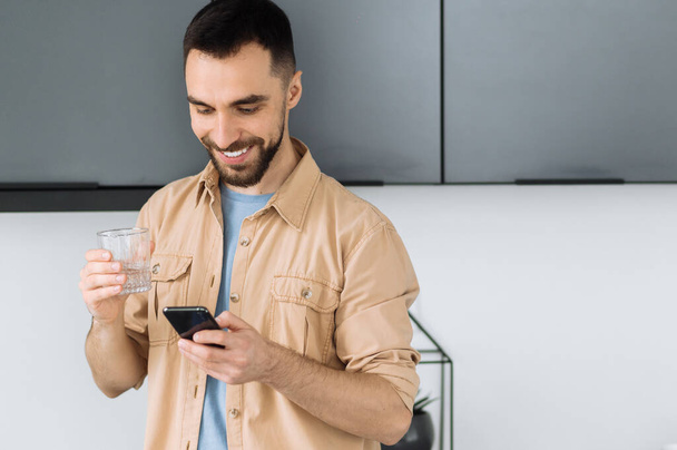 Brodaty biały mężczyzna używa smartfona, rozmawia ze znajomymi lub przegląda internet. Szczęśliwy człowiek zajęty trzymać szklankę czystej wody, przestrzegać zdrowego stylu życia i pić dzienną stawkę wody - Zdjęcie, obraz