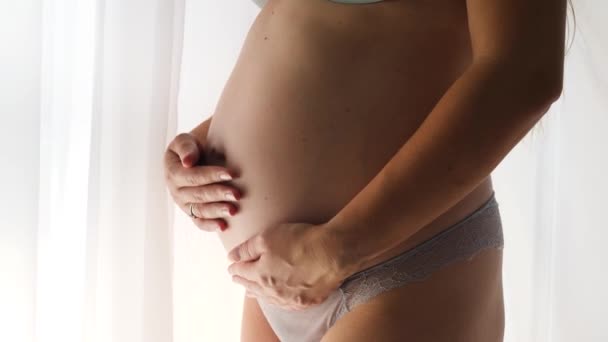 Close-up van een zwangere vrouw in lingerie die bij het raam staat en haar dikke buik vasthoudt. Concept van gelukkige zwangerschap en baby anticipatie - Video