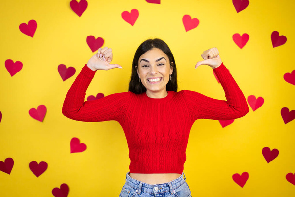 Junge kaukasische Frau auf gelbem Hintergrund mit roten Herzen, die selbstbewusst lächelnd auf sich selbst zeigen, mit stolzen und glücklichen Fingern. - Foto, Bild